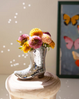 The Ceramic Disco Boot Vase by Kalalou, Inc gleams on a light wooden stool, filled with colorful flowers. Sunlight reflects off its mirrored surface while a blurred butterfly art piece adds charm—a unique gift for any cowgirl.