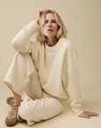 A woman with blonde hair sits on the floor against a beige background, wearing the Sold Out NYC Cashmere Signature Crew, cream wide-leg pants, and tan sneakers, looking directly at the camera with a relaxed expression.