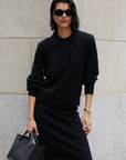 A woman in a Sold Out NYC Cashmere Polo and skirt, with black sunglasses, gold jewelry, and a black handbag stands against a stone wall.