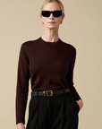 A blonde woman stands against a beige background wearing black sunglasses, gold hoop earrings, a dark brown Sold Out NYC Refined Crew sweater, and black high-waisted pants with a brown belt.
