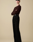 A woman confidently stands against a beige background wearing Sold Out NYC's Refined Crew sweater in dark brown, paired with high-waisted black wide-leg trousers, a belt, black shoes, and black sunglasses.