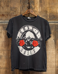 The Made Worn Guns N Roses Crew Tee, featuring two guns, two red roses, and the band’s name in a circular design, hangs on a wooden hanger against a rustic wood background—ideal for any Welcome to The Jungle fan.