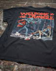 A black Guns N Roses Crew Tee by Made Worn rests on a paint-splattered surface. The back shows vivid skeleton and jungle artwork with bold red "WELCOME TO THE JUNGLE" text, inspired by Guns N Roses.