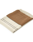 The Libeco Home Foundry Throw is a folded linen-wool blend with a beige base, wide brown stripe, thin black lines, fringed edges, and a stonewashed finish, shown on a white background.
