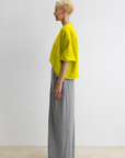 A person with short blond hair stands in profile against a white background, wearing the Rachel Comey Fond Sweatshirt in yellow and high-waisted, wide-leg gray pants.
