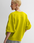 A person with short blonde hair, seen from behind, wears the Rachel Comey Fond Sweatshirt—a loose, bright yellow crewneck with wide cropped sleeves—paired with light gray pants against a plain light background.