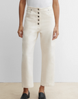 A person stands against a plain background wearing a white short-sleeve shirt, Rachel Comey Elkin Pant in Dirty Pomona Denim with button details and an adjustable back buckle, and black slip-on shoes.