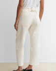 A person stands facing away, wearing Rachel Comey’s off-white Elkin Pant featuring an adjustable back buckle and back pockets, paired with a white t-shirt and black shoes, on a beige carpet against a plain white wall.