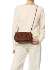 A person in a cream sweater and pants holds the Jerome Dreyfuss Bobi Bag, featuring brown leopard print goatskin and an adjustable chain strap, against a plain white background.