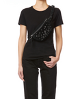 A person in a black t-shirt and black pants wears the Jerome Dreyfuss Lino Banana bag, a stylish cross-body decorated with silver grommets, slung across their chest. The background is plain white.