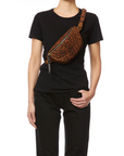 A person in a plain black t-shirt and pants models the Jerome Dreyfuss Lino Banana bag, featuring a brown leopard print, worn cross-body with one hand on their hip.