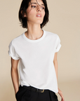 A person with short dark hair wears The Soft Perfect Tee from Sold Out NYC in organic Peruvian cotton and black pants, standing against a beige background and looking to the side with a relaxed expression.