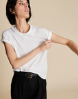 A person with short dark hair wears The Soft Perfect Tee by Sold Out NYC, a relaxed fit shirt made from organic Peruvian cotton, tucked into black belted trousers while adjusting their shirt against a neutral beige backdrop.