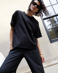 A person with short, wavy hair wearing black sunglasses, black pants, and The Oversized Tee by Sold Out NYC stands outdoors in front of a light stone building with a large window, leaning slightly forward.