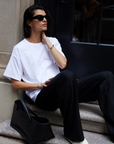 A person with shoulder-length dark hair and sunglasses sits on stone steps beside a black bag, wearing Sold Out NYC’s The Oversized Tee—crafted from buttery-soft heavyweight cotton—and black pants, resting a hand on their neck and looking aside.