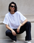 A woman with short dark hair and black sunglasses wears Sold Out NYC’s The Oversized Tee, black pants, and white sneakers as she squats confidently against a light stone wall.