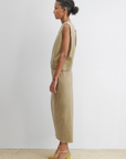 A woman stands in profile against a white wall, wearing the modern Rachel Comey Rini Dress—a sleeveless cotton style that falls below the knees—paired with yellow strappy high-heeled sandals. Her dark hair is pulled back into a bun.