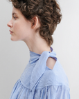 A person with short, curly brown hair is shown in profile wearing the Rachel Comey Parachute Top—a light blue, textured, wrinkle-resistant long-sleeve blouse featuring a high collar and bow detail—against a plain, light background.