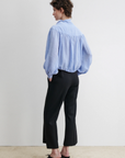 A person stands with hands in pockets, wearing the Rachel Comey Parachute Top— a loose light blue long-sleeve blouse— paired with black wide-leg trousers and heeled shoes, set against a plain white background.