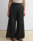 Someone stands against a plain background wearing Rachel Comey's Coxsone Pant—high-waisted, wide leg, black organic cotton—paired with a light short-sleeve top and brown open-toe sandals.