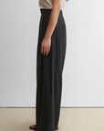 A person stands in profile against a plain background, wearing a short-sleeve beige top, brown flat sandals, and high-waisted wide leg Coxsone Pants by Rachel Comey in black.