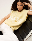 A woman with curly hair sits on a black leather chair, wearing the BA&SH Sweat Benta—a pale yellow sleeveless sweatshirt with American-style raglan armholes—paired with white pants and gold jewelry, looking directly at the camera with a serious expression.