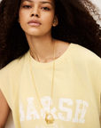 A young woman with voluminous curly hair wears the BA&SH Sweat Benta, a light yellow sleeveless sweatshirt with a round neckline and "BA&SH 2003 PARIS" print, accessorized with a long gold necklace, looking confidently at the camera.