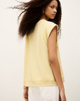 A woman with long curly hair models the BA&SH Sweat Benta, a sleeveless light yellow top with a round neckline, paired with white pants. She stands back to the camera, looking over her shoulder against a plain background.