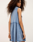 A person with long curly hair models the BA&SH Dress Ilona—a western-inspired, light blue denim dress—while looking over their shoulder against a plain backdrop.