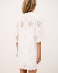 A woman with wavy blonde hair is seen from the back wearing the white, knee-length Dress Malo by BA&SH, featuring sheer lace patterns and elbow-length sleeves, set against a plain light background.