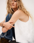 A woman with long wavy blonde hair wears the BA&SH Top Maxi— a sleeveless white broderie anglaise cami—paired with blue jeans, sitting with one knee up and looking at the camera against a plain background.