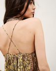 A woman with long dark hair is shown from the back, wearing the sleeveless Top Pamela by BA&SH. The top features thin, cross straps and an earthy-toned patterned fabric as she looks over her shoulder against a plain background.
