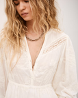 A woman with long, wavy blonde hair wears the BA&SH Dress Prisco with lace detail and delicate embroidery, accessorized with layered necklaces, looking down serenely against a soft, neutral background.