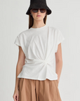 A woman in a black bucket hat, dark sunglasses, and loose tan pants models the Apiece Apart Volta Twist Top—an organic cotton tee with a twist detail at the waist—against a plain light background.
