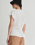 A person in the Apiece Apart Volta Twist Top, black bucket hat, sunglasses, and light brown shorts faces away from the camera against a plain background.