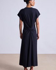 A woman stands facing away in the Apiece Apart Volta Twist Dress—a black organic cotton, ankle-length style with short sleeves and a gathered twist detail at the lower back. Her hair is in a bun, and she wears red strappy sandals against a plain background.