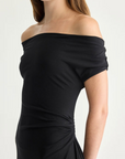 A woman poses against a plain light background wearing the Apiece Apart Marles Off the Shoulder Dress in black, featuring ruched waist detailing. She wears her brown hair down and accessorizes with geometric earrings.