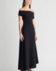 A woman wearing the Apiece Apart Marles Off the Shoulder Dress in black cotton jersey, paired with black sandals, a black headband, and silver jewelry, stands against a plain light background with a neutral expression.
