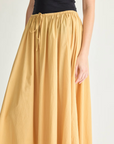 Against a plain light background, a person wears the Apiece Apart Hanne Tie Maxi Skirt—a long, flowing organic cotton skirt with a drawstring waist—paired with a black top. Only the torso and skirt are shown.