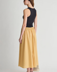A woman stands sideways against a plain light background, wearing a black sleeveless top, the Apiece Apart Hanne Tie Maxi Skirt in yellow, black sandals, sunglasses, and a black headband.