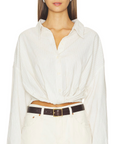 A woman with straight brown hair wears the Denimist TWISTED FRONT SHIRT in a relaxed fit, tucked into high-waisted white pants and cinched with a dark brown belt; the draped front of her shirt enhances her effortless style as she stands against a plain white background.