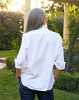 A person with long gray hair stands outdoors facing away in a relaxed-fit white button-down and Frank & Eileen Shirley Tattered Denim, exuding casual California style amid greenery and sunlight.