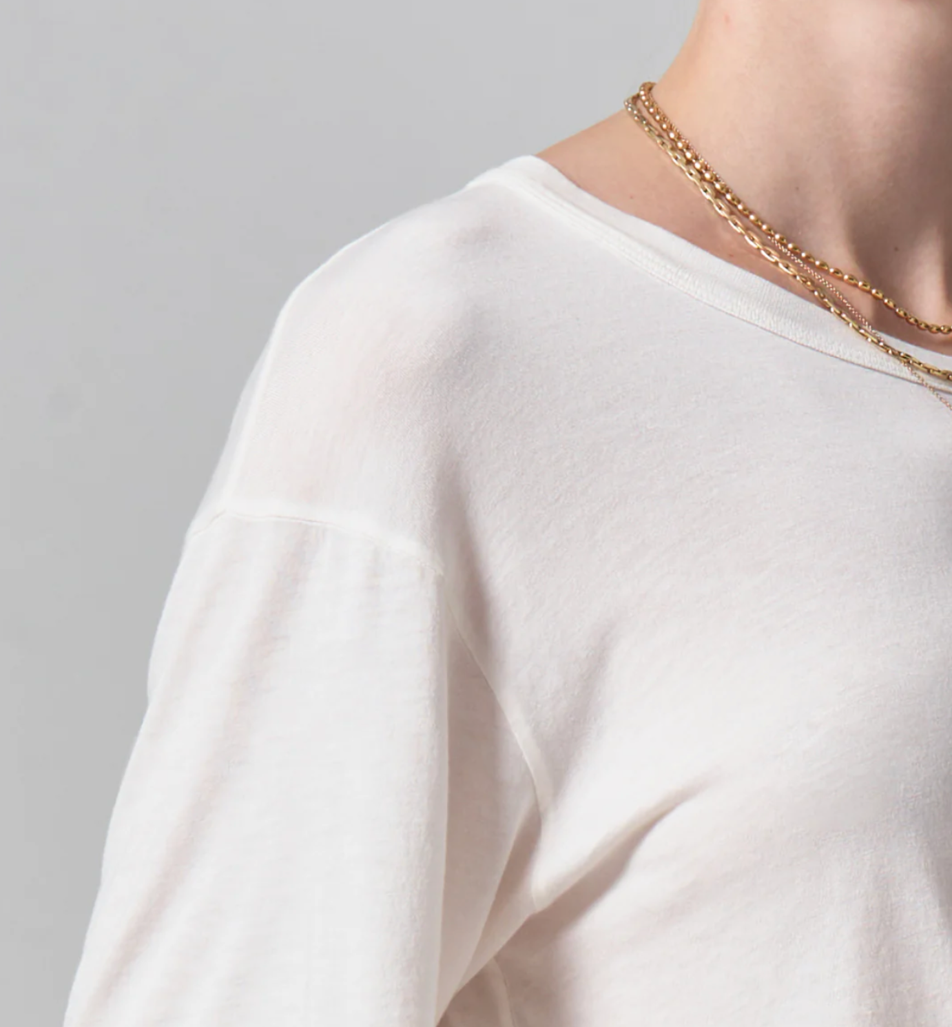 Close-up of a person wearing a Citizens Of Humanity/AGOLDE Elisabetta Relaxed Tee In Pashmina in Arizona style with gold necklaces, focused on the shoulder area against a gray background.