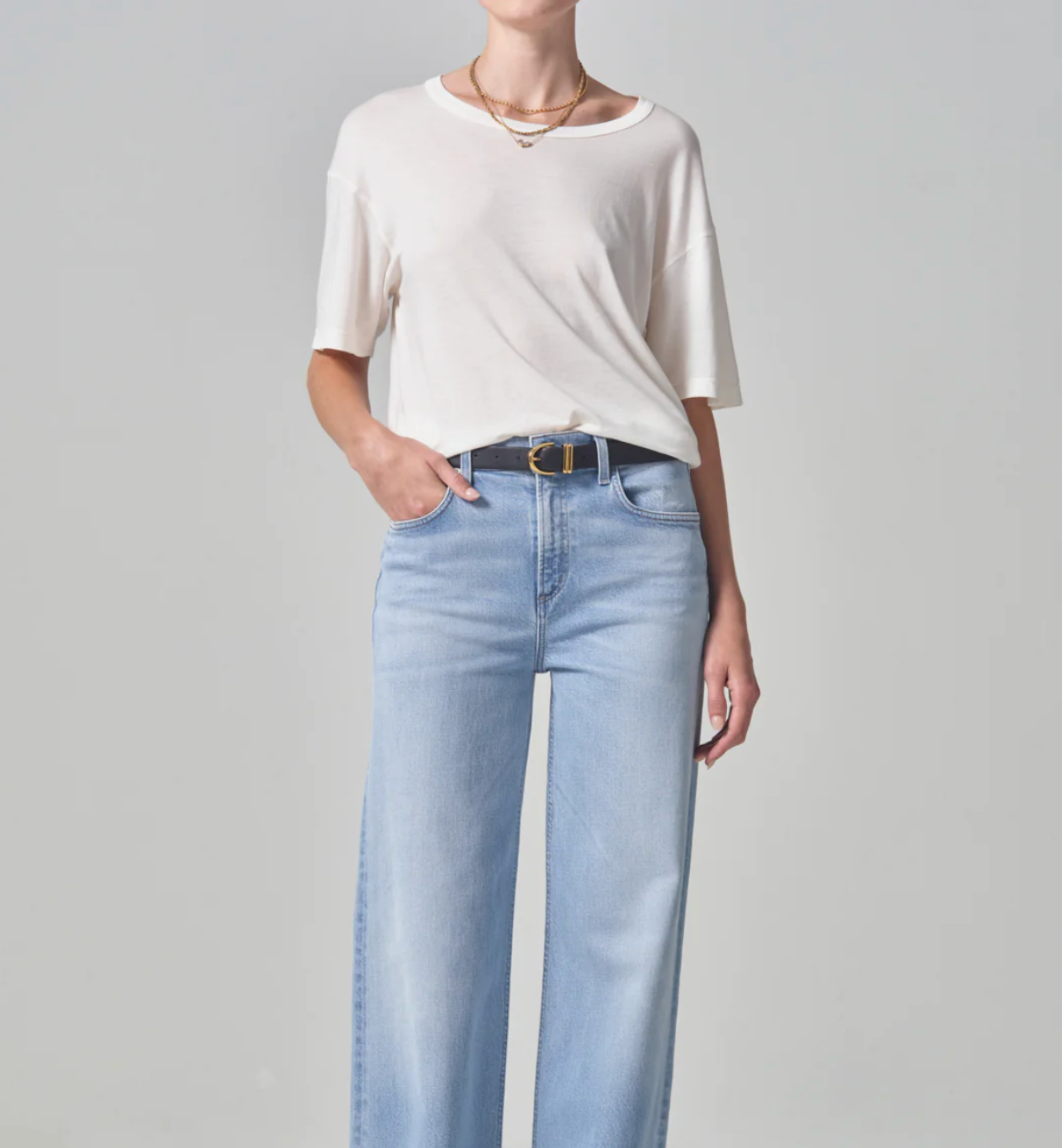 A woman in a simple white Elisabetta Relaxed Tee In Pashmina and blue jeans stands with one hand in her pocket against a plain background, focusing on Citizens Of Humanity/AGOLDE Arizona-style casual wear.