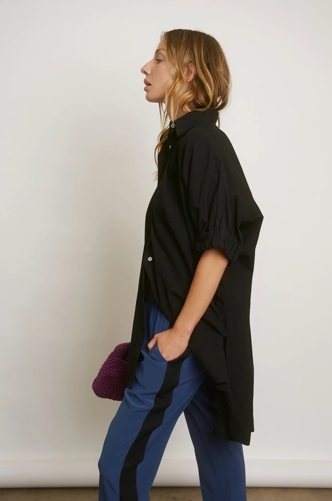 A woman in profile wears a black blouse and blue trousers, carrying a purple bag. She is standing against a plain light background, exuding Aquarius Cocktail style.
