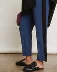 A person stands in a studio showing a style detail shot focused on the lower half of their body, wearing Aquarius Cocktail blue trousers with a black stripe, black loafers, and holding a purple mesh bag.