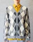 A person wears a yellow collared shirt under a gray, white, and black Brazeau Tricot Argyle Cardigan, standing against a light gray background. Only the upper body is visible.
