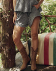 A person wearing The Delta Short by The Great Inc. and cowboy boots stands on a wooden porch near a rustic railing and a striped blanket draped over a bench.
