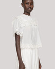 A woman wears the Leila Eyelet Top by Sea New York, a lightweight cotton blouse with eyelet lace and short sleeves, paired with a matching white skirt. She stands against a plain light gray background.