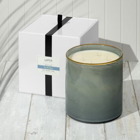 A large gray hand-blown glass vessel candle with three wicks, the Lafco NY Beach House Candle, sits on a white wooden surface beside its gift box tied with a black ribbon. Soft shadows fall across the scene.